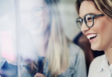 Lächelnde Frau mit Brille und längeren Haaren schaut auf einen Bildschirm.