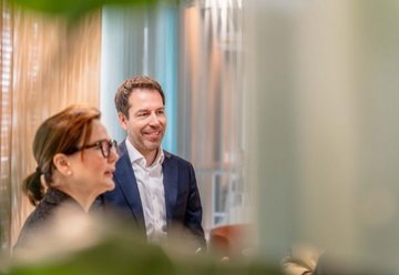 Zwei Personen in einem Büro, eine Frau im Vordergrund und ein lächelnder Mann im Anzug im Hintergrund.