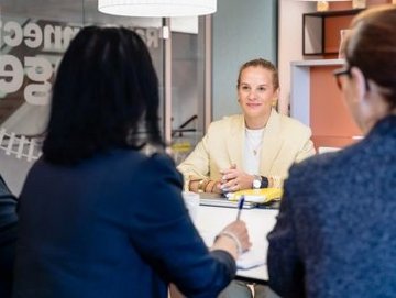Vier Frauen in einem Besprechungsraum; eine Frau im hellen Blazer sitzt gegenüber den anderen und wirkt aufmerksam und zugewandt.