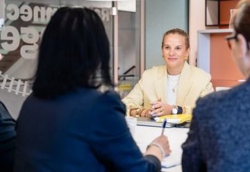 Vier Frauen in einem Besprechungsraum; eine Frau im hellen Blazer sitzt gegenüber den anderen und wirkt aufmerksam und zugewandt.