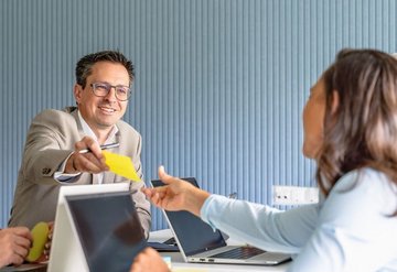 Vier Personen in einem Meeting; ein Mann reicht einer Frau einen gelben Zettel über den Tisch. Alle wirken freundlich und engagiert.