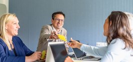 Vier Personen in einem Meeting; ein Mann reicht einer Frau einen gelben Zettel über den Tisch. Alle wirken freundlich und engagiert.