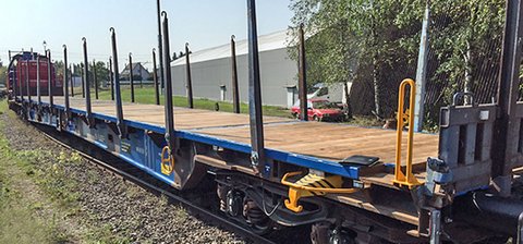 Blauer Flachwagen mit seitlichen Elementen vor einer grauen Halle.