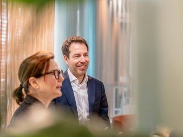 Zwei Personen in einem Büro, eine Frau im Vordergrund und ein lächelnder Mann im Anzug im Hintergrund.